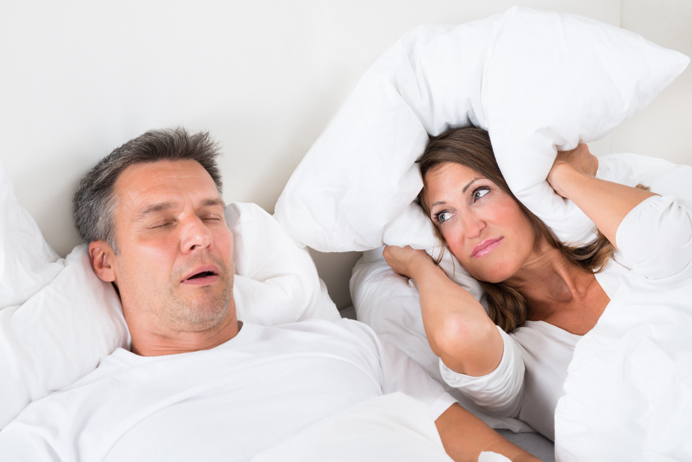 Man in bed snoring with woman covering her ears with pillow next to him